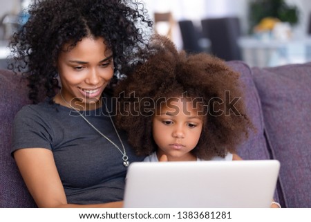 Smiling young African American mom relax with small daughter on couch at home with laptop watching cartoons online, millennial black mommy rest with little child, teach her using computer
