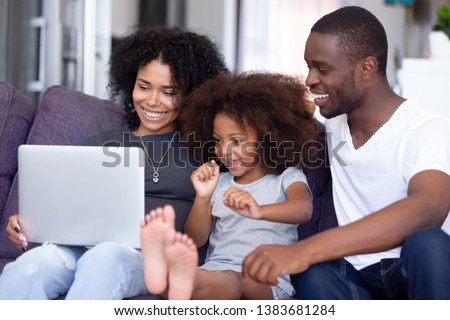 Excited young African American family rest on couch watching movie or game online cheering, happy child relax with millennial parents at home, have fun laughing enjoying cartoon on laptop