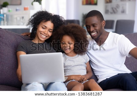 Happy African American young family relax on couch with laptop, watching movie or cartoon together, smiling mom, dad and little daughter have fun at home, using computer, resting on cozy sofa