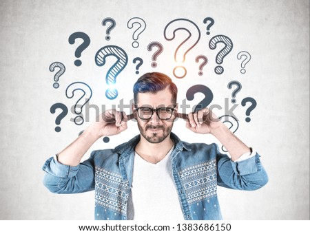Young man in glasses and casual clothes covering his ears standing near concrete wall with question marks drawn on it. Concept of ignoring problems.