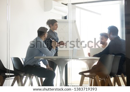 Millennial indian female speaker stand making flip chart presentation speak on company business strategy, diverse multiethnic work team brainstorm discuss share ideas meeting at briefing in office Royalty-Free Stock Photo #1383719393