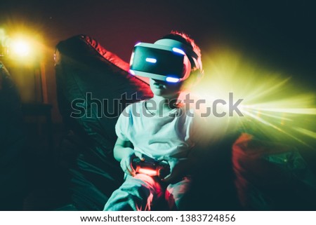 Little boy wearing VR goggles and playing game with gamepad in neon lights on black background on background bright projector illuminated boy.