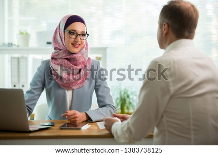 Coworkers meeting in office. Stylish woman in hijab making conversation at desk with man in white modern office. Muslim businesswoman in eyeglasses interviewing man.