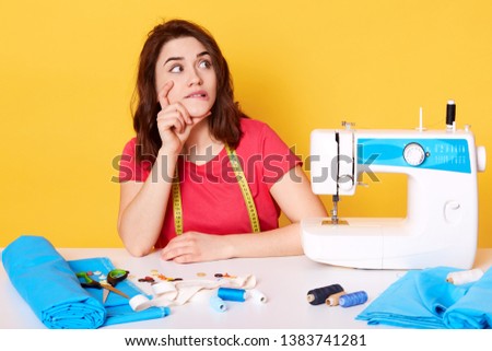 Thoughtful European young lady looks aside with interest, creating new ideas, having inspiration time, pause to sew. Modern sewing machine, thread and blue fabric are on table. Sewing concept.