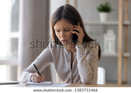Upset young woman sit at office desk talking over phone disappointed hear bad news, sad pensive girl writing take notes in notebook feel unhappy speaking having unpleasant cellphone conversation Royalty-Free Stock Photo #1383751466