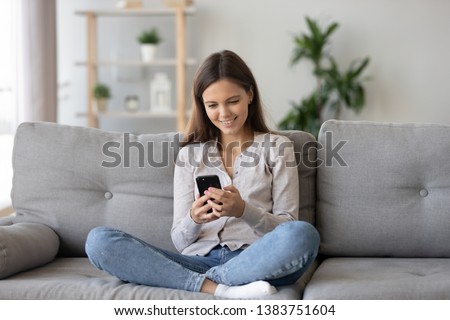 Smiling teenager girl relax sit on sofa at home using cellphone texting chatting with friend, happy young woman on couch hold smartphone shop online or check mobile application or social media Royalty-Free Stock Photo #1383751604