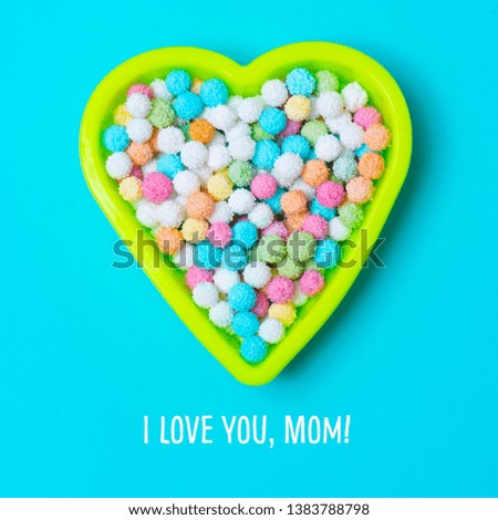 high angle view of a yellow plastic heart-shaped container full of candies of different colors and the text I love you mom on a blue background