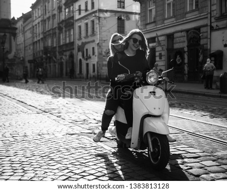 Young and beautiful girls in sunglasses riding a scooter. Portrait of pretty ladies riding a motorbike on old downtown street. Summer vacation. Girls in a black t-shirts on a vintage scooter