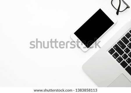 White smartphone with black blank mock up screen is on top of white office desk table with laptop computer. Top view with copy space, flat lay.