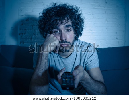 Young bored man on couch using TV remote control zapping for another movie or show late at night. Looking disinterested and sleepless. In entertainment People insomnia and Sedentary lifestyle concept. Royalty-Free Stock Photo #1383885212