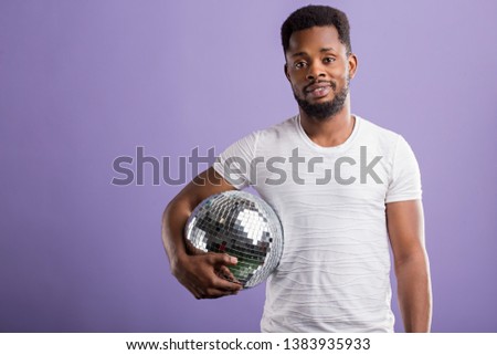 Happy dark skinned man holding shining disco ball, going to party with friends, celebrating something, wearing casual clothes, smiling standing over purple background with copy space.