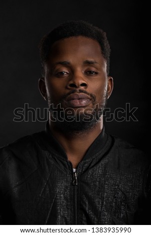 Highly detailed closeup portrait of attractive young african man wearing black jacket looking at camera with serious and thoughtful expression on dark background. Human emotions and feelings.
