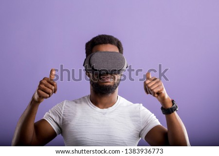 Dark skinned young man experiencing virtual reality, showing thumb up, playing video games using oculus rift headset. 3d technology, innovation, entertainment, cyberspace concept.