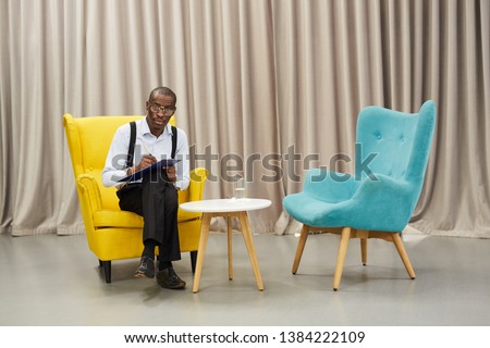 Full length portrait of African-American man holding clipboard  posing looking at camera while  sitting on  design chair against drapery