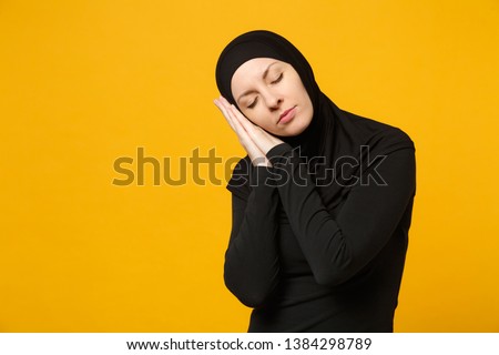 Image beautiful young arabian muslim woman in hijab black clothes sleep with folded hands under cheek isolated on yellow wall background. People religious Islam lifestyle concept. Mock up copy space.