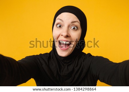 Close up young arabian muslim woman in hijab black clothes doing selfie shot on mobile phone isolated on yellow wall background studio portrait. People religious lifestyle concept. Mock up copy space