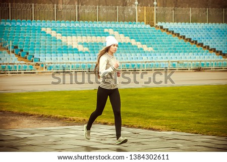 
A young girl with long hair in a white knitted hat, black leggings and a sports jacket, sneakers runs against the backdrop of a football net, green grass and sports stands.