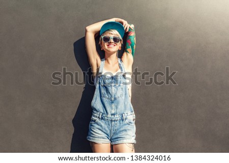 Young alternative girl wearing cap and sunglasses standing isolated on grey wall on the city street hands up relaxed smiling cheerful