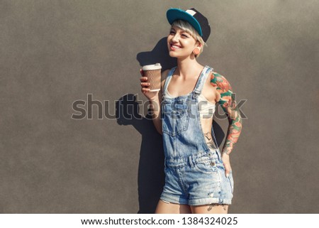 Young alternative girl wearing cap standing isolated on grey wall on the city street holding cup drinking hot coffee looking aside smiling cheerful