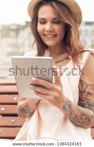 Young woman wearing hat sitting on bench outdoors on the city street holding digital tablet close-up using application laughing cheerful