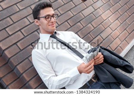 Young business man standing leaning on wall wearing eyeglasses on the city street counting dollar banknotes smiling happy