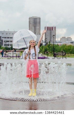 Young wet pretty girl with two braids in yellow boots and with transparent umbrella stands inside of fountain. Rainy day in city.