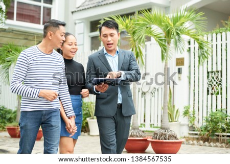 Asian young realtor showing photos of new house to young family on tablet pc while they walking along the street
