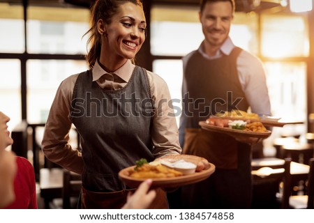 Two waiters serving lunch and brining food to their gusts in a tavern. Focus is on happy waitress.  Royalty-Free Stock Photo #1384574858