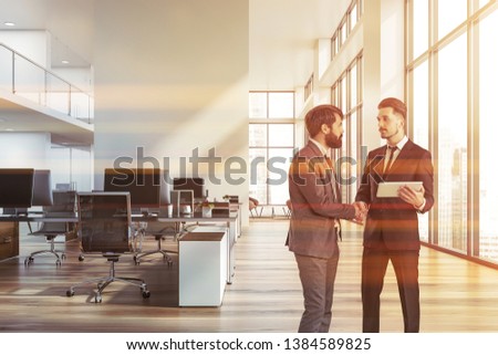 Two young businessmen shaking hands in open space office with rows of computer tables and panoramic windows. Concept of agreement. Toned image