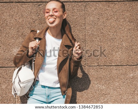 Portrait of beautiful smiling brunette model dressed in summer hipster jacket clothes. Trendy girl posing in the street near wall. Funny and positive woman in round sunglasses. Showing her tongue