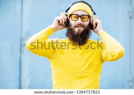 Portrait of a stylish bearded man in yellow clothes enjoying music with headphones on the blue background