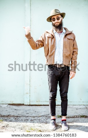 Full body portrait of a stylish bearded man dressed in jacket and hat on the light turquoise background outdoors