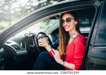 Beautiful young cheerful woman in fashion sunglasses with coffee in hand sitting in black car. Female on road trip drinking coffee inside car. Lifestyle People Travel concepts
