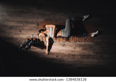 Top view of young Caucasian brunette lying down on mat and stretching in gym at night. Next to her jumps footwear.