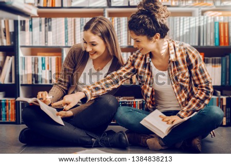 Two Caucasian pretty female students sitting with legs crossed in library and studying for exams. Girl with curly hair pointing at answer in book.