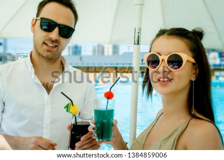 Young beautiful couple, on the background of the pool, on vacation, with cocktails in hand, looking at the frame and smiling. Summer vacation concept