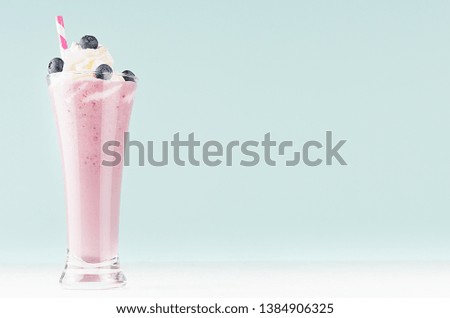 Juicy violet smoothie of blueberry with berries, striped straw, whipped cream on white wood board and light pastel blue background, copy space.