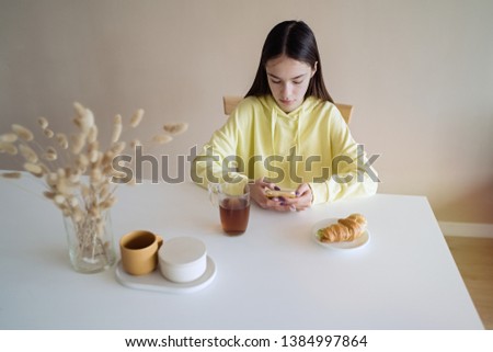 Cute teen girl eating breakfast in kitchen while using on her smart phone                              