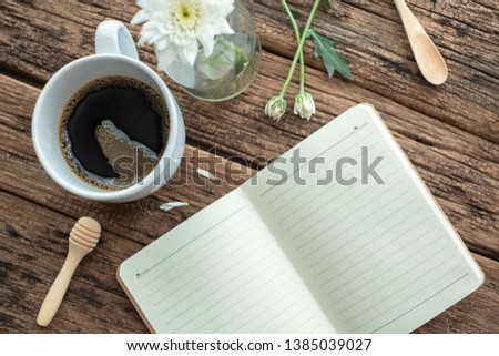 Black coffee cup with white flower& blank notebook on wood table. The morning light from window makes the house more cozy& romantic. This photo is good for chillout music cover. Lazy Sunday concept. 