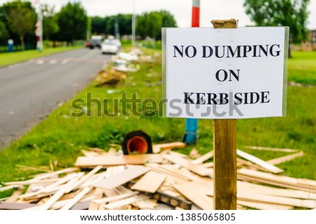 A sign on a grass verge warning people not to dump rubbish on the kerb side, with a lot of discarded wood, rubbish and other items littering the ground.