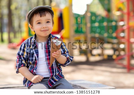 Portrait of a happy toddler with dreamy face expression sitting in checkered shirt and flat cap on a bench in park with ice cream in waffle cone in sunny spring or summer day. Playground in background