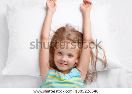 Top view of pretty child has long hair, stretches in bed, lies on white pillow, has good mood after rest in bed, awakes early in morning, wants to start new day, looks directly at camera. Childhood