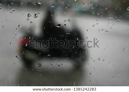 Rain drops on a wet window with an intentionally blurred city scene in the background featuring a person riding on a motorbike. Concept is of gloomy, rainy weather in a large city. 