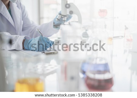 A scientist hands writing on a clipboard in laboratory with test tube microscope and solutions. Royalty-Free Stock Photo #1385338208