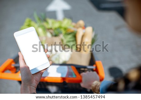 Holding smart phone with empty screen with shopping cart full of food on the background. Online shopping concept