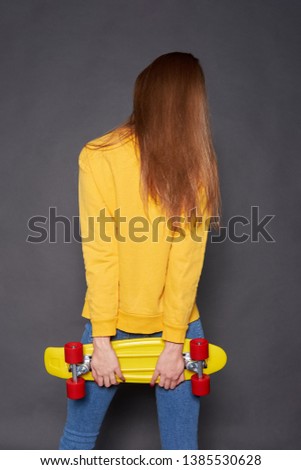 Stylish redhead girl wearing yellow sweatshirt and blue jeans holding yellow skateboard with red wheels. Studio shot on grey background. View from back. 