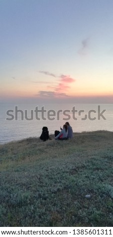 Romantic beautiful background. Young lovers man and woman on a cliff of the sea against the sunset sky. Next to people is a big dog. People taking pictures of the sea sunset. Summer evening