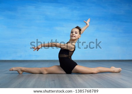 A girl gymnast in a black gymnastic swimsuit is sitting on a cross splits on the blue sky background.