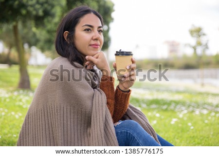 Tranquil pretty girl enjoying morning outdoors. Young woman in warm clothes sitting on grass, drinking takeaway coffee and looking into distance. Morning outdoors concept