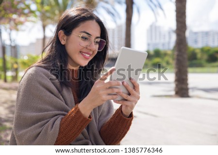 Smiling pretty woman using tablet computer outdoors. Young woman holding gadget with blurred trees and walkway in background. Technology concept.